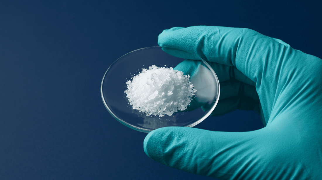 Nahaufnahme einer kristallinen Chemikalienprobe auf einem Uhrglas, gehalten mit einem Nitrilhandschuh.