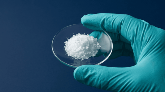 Nahaufnahme einer kristallinen Chemikalienprobe auf einem Uhrglas, gehalten mit einem Nitrilhandschuh.